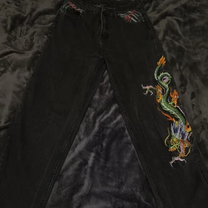 Svarta Ed Hardy jeans med drakbroderi - Unika svarta jeans från Ed Hardy med färgstarkt broderad drake längs ena benet. Klassisk femficksmodell med coola mönstrade detaljer i fickorna och insidan. Jeansen har dragkedja och knapp med ED-logga. Perfekt för dig som vill sticka ut med edgy streetstyle.