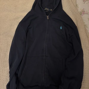 Marinblå hoodie från Polo Ralph Lauren - Säljer en marinblå hoodie från Polo Ralph Lauren med dragkedja och huva. Tröjan har klassisk broderad logga i ljusblått på bröstet och stora fickor framtill. Perfekt för chill dagar och enkel att matcha med jeans eller joggers. Snören saknas därför är priset så lågt!