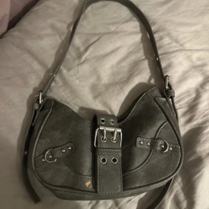Vintage grå axelväska med bältdetalj - Snygg grå axelväska i syntetmaterial med coola bältdetaljer och silvriga nitar. Väskan har en böjd form och justerbar axelrem. Perfekt för dig som gillar en edgy stil och vill ha en unik accessoar till din outfit. Lite stilage men det är en självklarhet då väskan är vintage. 