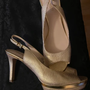 Guldiga pumps med öppen tå och klack - Snygga pumps från Nine West i skimrande guld med öppen tå och hög klack. Skorna har en elegant plattform och justerbar rem runt hälen. Materialet har en glittrig finish som ger extra lyxig känsla. Perfekta för dig som vill sticka ut med stil. Använda med små märken/normal sliten