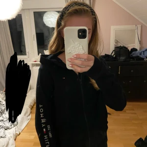 Svart zip hoodie från Tommy Hilfiger - Oanvänd svart zip hoodie från Tommy Hilfiger med dragkedja och huva. Storlek 164 men passar xs-s. Snygga vita och röda detaljer på kragen och logotyp på bröstet samt tryck längs ena ärmen. Nypris runt 700 kr. Säljer då den inte används.❣️