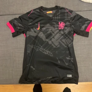 Svart Nike fotbollströja med rosa detaljer - Snygg svart fotbollströja från Nike med grått grafiskt mönster och rosa detaljer. Tröjan har korta ärmar, V-ringning och ett broderat rosa lejon på bröstet. Materialet är lätt och andas, perfekt för träning eller match. Swoosh-loggan i rosa och gult ger extra stil.
