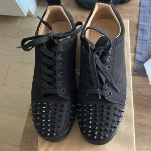 Christian Louboutin svarta sneakers med nitar - Svarta sneakers från Christian Louboutin i mocka med svarta nitar på tån och klassisk röd sula. Skorna har snörning och en stilren, låg profil. Perfekta för dig som vill sticka ut med en edgy och lyxig vibe.