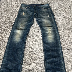 Blå jeans med slitningar, straight fit - Säljer ett par blå jeans med coola slitningar och ljusa tvättade partier på låren och knäna. Modellen har klassisk femficksdesign och rak passform som sitter snyggt på benen. Perfekt för dig som gillar en avslappnad streetstil.