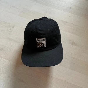 Okey keps - Säljer en svart keps från Obey med klassisk böjd skärm och justerbar snapback baktill. Framtill sitter en broderad vit Obey-logga med grafiskt motiv. Kepsen är tillverkad i bomull och har en stilren, enkel design som passar till streetwear. Knappt använd, undanlagt bland andra kläder och därmed blivit lite skrynklig, vilket ej syns när man väl tar på sig den