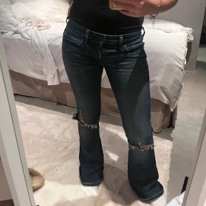 Mörkblå bootcut jeans från Hollister - Snygga mörkblå jeans från Hollister med bootcut passform och slitna detaljer vid knäna. Klassisk femficksmodell med hög midja och tydliga sömmar. Jeansen är tillverkade i stretchigt denim för skön passform och har en cool, lite edgy vibe.