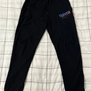 Svarta mjukisbyxor från Trapstar - Svarta mjukisbyxor från Trapstar med färgglad logga på vänster höft. Byxorna har snörning i midjan och är tillverkade i 100% bomull. Perfekta för chill eller streetwear, med en loose passform och bekväm känsla.