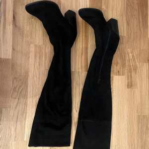 Svarta overknee stövlar i mocka - Säljer ett par svarta overknee stövlar med spetsig tå och bred klack. Skorna är gjorda i mjuk mocka och har dragkedja på insidan för enkel påtagning. Perfekta för att ge din outfit en edgy vibe och passar till både jeans och kjol.