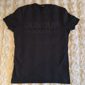 Svart t-shirt från Dondup - Svart t-shirt från Dondup med diskret tryck på bröstet. Klassisk rund hals och korta ärmar. Materialet känns mjukt och skönt, perfekt för en clean och stilren look. Made in Italy står på etiketten, vilket ger extra premiumkänsla.