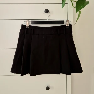 Svart kortkjol från MNG, strl 34 - Snygg svart kortkjol från MNG med klassisk A-linjeform och breda bälteshällor. Kjolen är ca 30 cm lång och har stilrena veck framtill. Perfekt för dig som gillar minimalistisk och clean stil. Materialet är mjukt och känns som bomull. Knapt använd. 