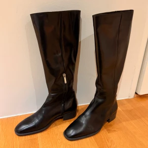 Svarta höga stövlar i skinn Zara - Snygga svarta stövlar från Zara med platt sula och rund tå. Tillverkade i slätt skinn med diskret dragkedja på sidan. Klassisk och stilren modell som passar till många olika outfits. Mycket bra skick då de knappt är använda och storleken 38.