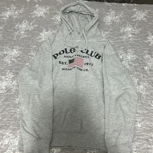 Grå tracksuit Polo Club Saint Vincent - Grå tracksuit från Polo Club Saint Vincent med tryck av amerikansk flagga och text på bröstet. Tröjan har huva med snörning och långa ärmar. Perfekt för chill dagar och snygg streetstyle.