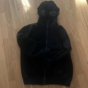 Svart hoodie med goggles från C.P. Company - Svart hoodie från C.P. Company med ikonisk huva som har inbyggda goggles och dragkedja hela vägen upp. Tröjan har en loose passform och är tillverkad i mjuk bomull. Perfekt för dig som vill sticka ut med streetwear-vibe.