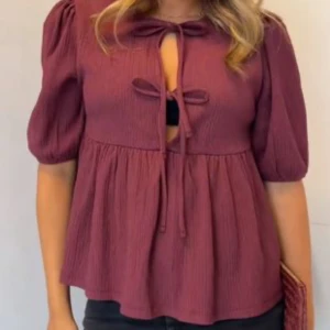 Vinröd blus med knyt och puffärm - Vinröd blus med puffiga halvlånga ärmar och två knyt framtill som skapar en snygg öppning. Blusen har en lös passform och faller fint över kroppen. Perfekt för dig som gillar trendiga detaljer och en feminin look.