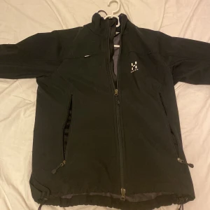Svart vindjacka från Haglöfs - Svart vindjacka från Haglöfs med Windstopper-membran. Jackan är tillverkad i 100% polyester, har hög krage, två dragkedjeförsedda fickor och justerbar nederkant. Perfekt för blåsiga dagar och outdoor-aktiviteter.