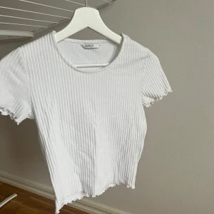 Vit ribbad topp från ONLY - Säljer en vit ribbad topp från ONLY med korta ärmar och rund hals. Toppen har vågiga kanter vid ärmslut och nederkant som ger en söt touch. Perfekt för dig som gillar enkel och clean stil med lite extra detalj.