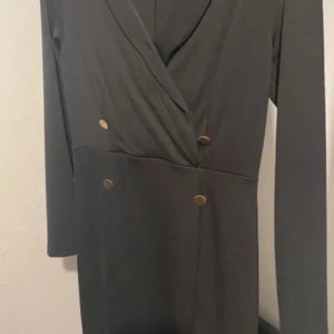 Svart omlottklänning med guldknappar - Stilren svart omlottklänning med långa ärmar och dekorativa guldknappar framtill. Klänningen har en v-ringad omlottfront som ger en elegant siluett. Perfekt för dig som gillar klassiska detaljer och enkel design.