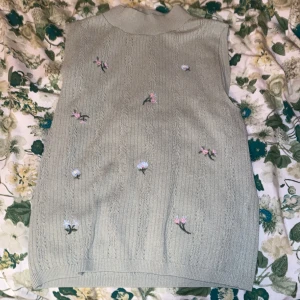 Stickad blommig väst från Zara - Supersöt ljusgrön stickad väst från Zara med små broderade blommor i pastellfärger. Västen har en mjuk ribbad krage och diskret mönsterstickning som ger en vintagekänsla. Perfekt att styla över en skjorta eller t-shirt för en trendig look.