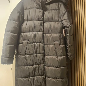 Svart lång pufferjacka i polyester - Lång svart pufferjacka med huva och stora fickor. Jackan är quiltad och har tryckknappar framtill samt på ärmarna. Tillverkad i 100% polyester, både yttertyg och vaddering. Perfekt för kalla dagar när du vill hålla dig varm och stilsäker.