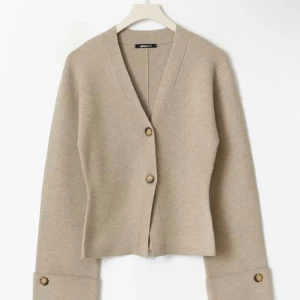 Beige kofta från Gina Tricot - Snygg beige kofta från Gina Tricot med v-ringad hals och två knappar framtill. Koftan har långa ärmar med breda muddar och dekorativa knappar vid ärmslutet. Perfekt att slänga på sig när det är lite kyligt ute.