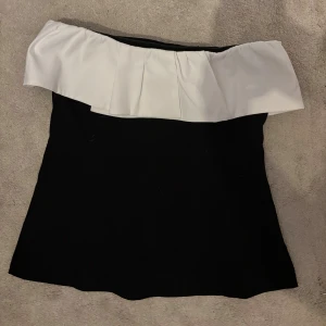 Svart och vit axelbandslös tubtopp - Säljer en svart tubtopp med bred vit volang upptill som ger en snygg kontrast. Toppen är axelbandslös och har en stilren, elegant look som passar perfekt till finare outfits.