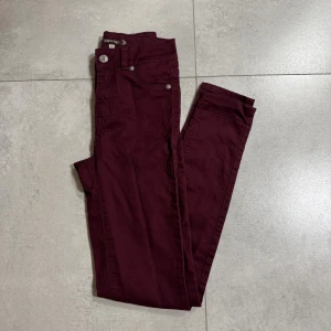 Vinröda skinny byxor Now and Then XS - Snygga vinröda byxor från Now and Then i storlek XS. Modellen är skinny med hög midja och klassiska fem fickor. Byxorna har dragkedja och knapp framtill och är tillverkade i mjuk bomull med lite stretch för skön passform.