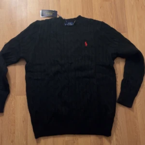 Svart kabelstickad tröja Polo Ralph Lauren - Svart kabelstickad tröja från Polo Ralph Lauren i storlek M. Tröjan har rund hals, lång ärm och den klassiska röda broderade loggan på bröstet. Materialet är mjuk ull som känns riktigt skönt mot huden. Perfekt för dig som gillar stilrena och tidlösa plagg.