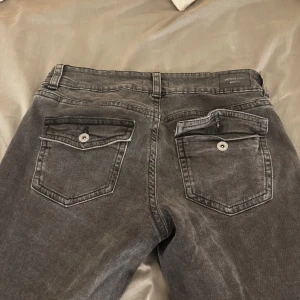 lågmidjade y2k jeans - gråa figurnära bootcut low waist jeans från Gina tricots kollektion ” perfect jeans” i storlek 40. Dom är knappt använda och har inga defekter. Bakfickorna har lock och knapp.  Enligt Gina Tricots hemsida: Midja: 75 cm Stuss: 103 cm Innerbenslängd:80 cm  Nypris: 500 kr