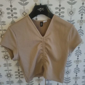 Beige ribbad croppad topp från Divided - Säljer en beige croppad topp från Divided med ribbad struktur och snygg snörning framtill. Toppen har korta ärmar och v-ringad hals, perfekt för dig som gillar trendiga och bekväma plagg. Passar dig som vill ha en stilren och cool look.