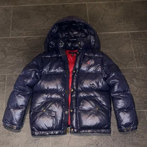 Mörkblå vinterjacka från Polo Ralph Lauren - Säljer en mörkblå vinterjacka från Polo Ralph Lauren med röd insida och huva. Jackan har guldiga knappar, dragkedja och två stora fickor framtill. Klassisk pufferstil med broderad logga på bröstet. Perfekt för kalla dagar. Hör av er vid minst lilla fråga eller fundering! 