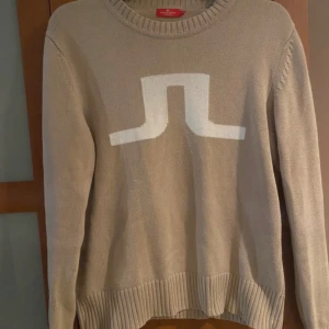 Beige stickad tröja från J.Lindeberg - Snygg beige stickad tröja från J.Lindeberg med deras ikoniska vita logga framtill. Tröjan har rund halsringning, ribbade muddar och är långärmad. Perfekt för dig som gillar stilrena och klassiska plagg med en modern touch.