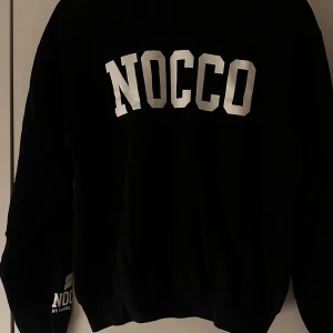 Svart NOCCO sweatshirt - Svart sweatshirt från NOCCO med stor vit logga på ryggen och tryck på ärmen. Klassisk passform med rund hals och långa ärmar. Perfekt för dig som gillar sportig och clean stil. 