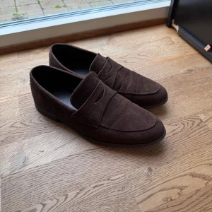 Bruna mockaloafers från Pier One - Snygga bruna loafers från Pier One i mjuk mocka med klassisk pennyloafers-design. Skorna har rund tå och platt sula, perfekt för en stilren look. Passar dig som gillar tidlös och clean stil med en touch av elegans.