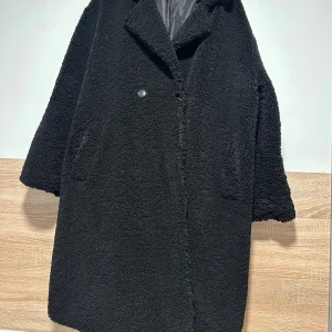 Svart teddyjacka med knappar - Mysig svart teddyjacka med oversized passform och stora slag. Jackan har två fickor framtill, knäppning med knapp och är helfodrad. Perfekt för kalla dagar när du vill vara både varm och stilren. Använd ett fåtal gånger, som ny.