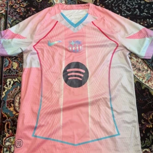 Rosa FC Barcelona Nike fotbollströja - Snygg FC Barcelona fotbollströja från Nike i ljusrosa och turkosa toner med vita ränder. Kortärmad med Dri-Fit material som andas. Tröjan har klubbmärke, Nike-logga och Spotify sponsortryck på bröstet. Modern design med V-ringad krage och grafiska detaljer.