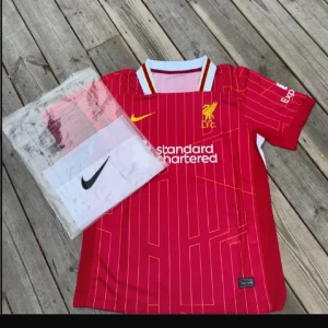 Liverpool FC röd matchtröja Nike - Liverpool FCs officiella matchtröja i rött med gula detaljer och tunna gula ränder. Tröjan har vit och gul krage, klubbmärke och Nike-logga på bröstet samt sponsortryck. Tillverkad i lätt och ventilerande material, perfekt för fotbollsträning eller match.