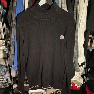 Svart polotröja från Moncler, L - Svart polotröja från Moncler i storlek L. Tröjan är ribbstickad med hög krage och har Moncler-loggan broderad på bröstet. Nederkanten har en snygg detalj med röd, vit och blå rand. Perfekt för dig som gillar stilrena och exklusiva plagg.
