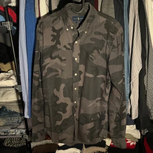 Camouflage skjorta från Ralph Lauren - Snygg långärmad skjorta från Ralph Lauren med mörkgrått camouflage-mönster. Slim fit-modell med klassisk krage och knappar hela vägen framtill. Tillverkad i mjuk bomull som känns skön mot huden. Perfekt för dig som gillar streetstyle och vill sticka ut.