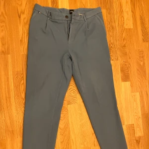 Blå kostymbyxor från BOSS - Snygga blå kostymbyxor från BOSS med klassisk rak passform. Byxorna har bälteshällor, knapp och dragkedja framtill samt fickor både fram och bak. Perfekta för en clean och stilren look. Materialet känns mjukt och har en lätt struktur.