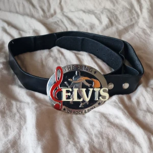 Svart Elvis skärp med stor buckle - Svart bälte i äkta skinn från Star Fashion med en stor, detaljerad buckle där det står 'ELVIS - The King of Rock & Roll'. Bucklet har inslag av rött, silver och orange, samt en siluett av Elvis. Perfekt för dig som gillar musik och vill sticka ut. Det har gjorts ett extra hål i bältet och tyget och som suttit fast på lädret har börjat släppa. Ca 90 cm från spännet till mittenhålet.