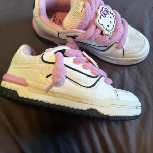 Hello Kitty sneakers med fluffiga snören - Säljer ett par vita sneakers med rosa fluffiga snören och Hello Kitty-detaljer på tungan och snörningen. Skorna har svarta och rosa detaljer, rund tå och platt sula. Ovandelen är i syntet och mesh för extra komfort. Perfekta för dig som älskar kawaii-stil!