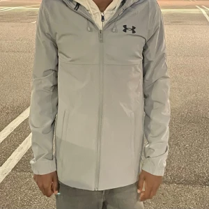 Ljusgrå vindjacka från Under Armour - Snygg ljusgrå vindjacka från Under Armour med huva och dragkedja framtill. Jackan har logga på bröstet och stort Under Armour-tryck på ryggen. Perfekt för dig som gillar sportig stil och vill ha en lätt jacka med clean look. Den helt ny också 