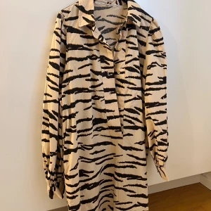 Zebramönstrad skjorta Second Female - Beige skjorta från Second Female med svart zebramönster och guldfärgade knappar. Skjortan har klassisk krage, långa ärmar med knäppning vid mudden och loose passform. Materialet är mjukt och lätt, perfekt för dig som gillar att sticka ut.