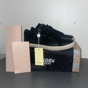 Golden Goose svart sneakers stjärna - Svarta sneakers från Golden Goose med klassisk stjärndetalj på sidan, snörning och vit platt sula. Skorna är i skinn med svarta mockadetaljer och har en stilren, minimalistisk look. Perfekta för dig som gillar streetstyle och vill ha något som sticker ut.