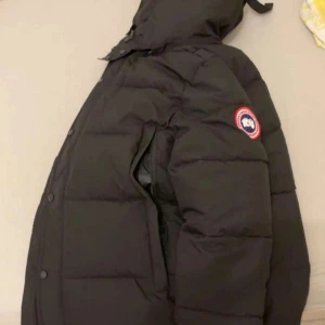 Svart dunjacka från Canada Goose - Svart dunjacka från Canada Goose med huva och avtagbar pälsdetalj. Jackan har klassisk patch på ärmen, dragkedja och knappar framtill. Fylld med dun och har en loose passform, perfekt för kalla vinterdagar. Snygg och tidlös design som håller dig varm. Storlek M och kommer med prislapparna.