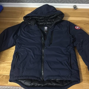 Mörkblå dunjacka från Canada Goose - Snygg mörkblå dunjacka från Canada Goose med huva och klassisk logga på ärmen. Jackan har dragkedja framtill, flera fickor med dragkedja och quiltad insida. Perfekt för kalla dagar och riktigt skön passform.