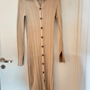 Beige ribbad långklänning med knappar - Långklänning i beige ribbat material med långa ärmar och knappar hela vägen framtill. Klänningen har en v-ringad krage och en smickrande, figurnära passform. Perfekt för dig som gillar stilrena och enkla plagg med snygga detaljer.