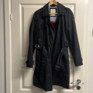 Svart trenchcoat från Esprit med bälte - Svart trenchcoat från Esprit med klassisk krage, dubbelknäppning och bälte i midjan. Jackan har långa ärmar, snygga detaljer vid axlar och ärmslut samt ett ljust mönstrat innerfoder med röd kant. Perfekt för dig som gillar stilrena och tidlösa plagg.
