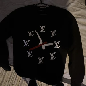 Svart Louis Vuitton sweatshirt med klocktryck - Svart sweatshirt från Louis Vuitton med stort klockinspirerat tryck på bröstet, där LV-loggor markerar timmarna. Tröjan har rund hals, långa ärmar och är tillverkad i mjuk bomull. Perfekt för dig som vill sticka ut med en lyxig och unik look. Tyvärr så är det ett pytte litet hål men det syns inte när du har på dig den 