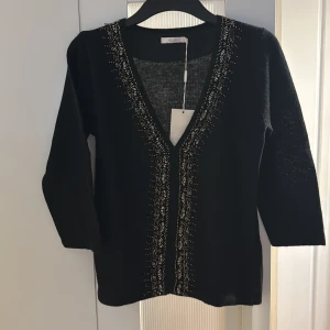 Svart kofta med pärldetaljer från Wera - Svart kofta från Wera med snygga pärldetaljer längs v-ringningen. Koftan har trekvartsärm och är tillverkad i en lyxig mix av lammull och angora, vilket gör den supermjuk och varm. Perfekt för dig som vill ha något stilrent men ändå med det lilla extra.